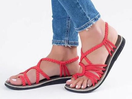Une personne porte un jean bleu à ourlet effiloché et des Sandales de Plage Colorées Olivia's Boutique à lanières tressées rouges, stylisées pour un look estival. Les sandales sont dotées de semelles plates et foncées offrant un confort exceptionnel. Leurs ongles sont peints en blanc sur un fond blanc uni dans la vue de côté.