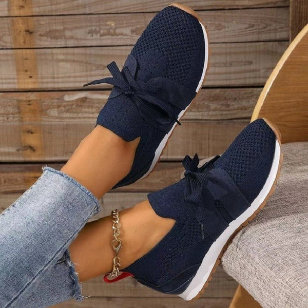 Une personne en jean bleu clair et une chaîne de cheville sur le pied droit porte les Chaussures Orthopédiques Anti-Douleur dOlivias Boutique, avec un tricot bleu foncé avec des semelles blanches et un détail de nœud, reposant avec style sur une surface en bois pour un soutien confortable.