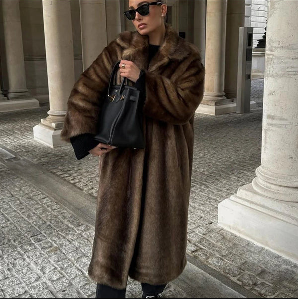 Dans une colonnade extérieure, une personne portant le luxueux Manteau long en fausse fourrure pour femme dOlivias Boutique en marron riche et lunettes de soleil foncées se tient gracieusement, tenant un sac à main noir qui respire lélégance et le style sur fond de colonnes de pierre et dun chemin pavé.