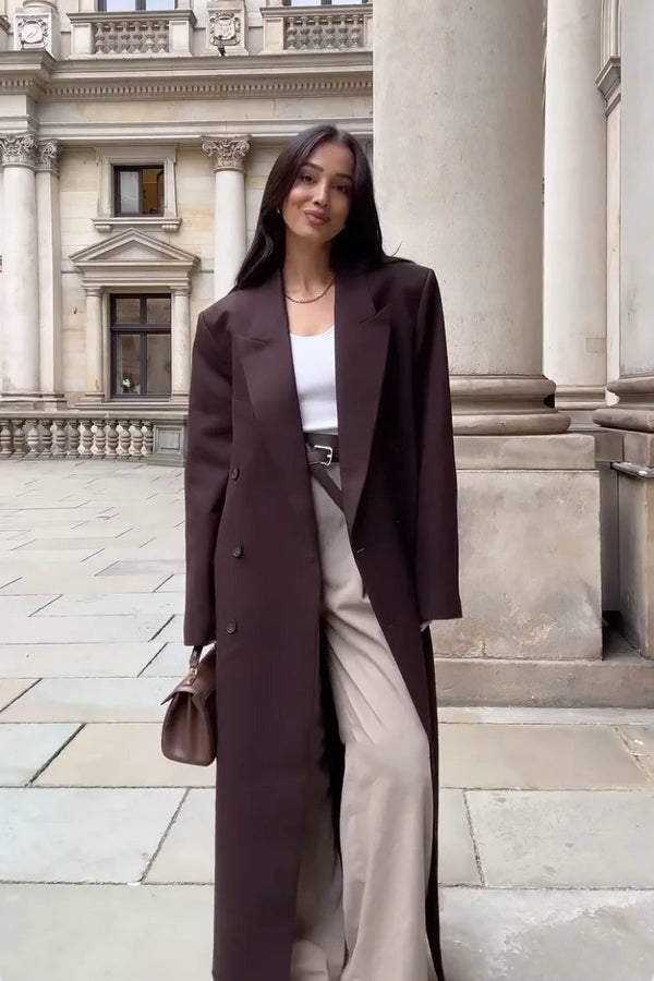 Une femme vêtue d'un manteau long uni et d'un pantalon beige d'Olivia's Boutique se tient debout sur une passerelle en pierre, dégageant une élégance classique alors qu'elle tient un sac à main marron. Le décor d'architecture historique avec de hautes colonnes et des pierres travaillées complète parfaitement son style intemporel.