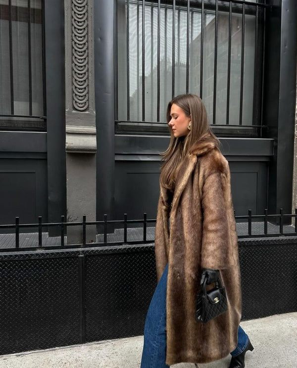 Vêtue du luxueux Manteau long en fausse fourrure pour femme dOlivia Boutique, une personne déambule sur un trottoir de la ville avec un sac à main noir et un jean bleu. De grands immeubles aux fenêtres et aux balustrades métalliques forment larrière-plan.