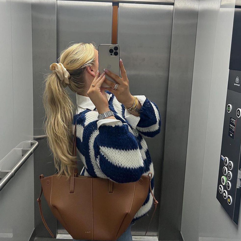 Dans un ascenseur, une personne prend un selfie devant un miroir, capturant ses longs cheveux blonds attachés avec un chouchou. Elle porte un pull rayé élégant et confortable pour toutes les saisons dOlivias Boutique et porte un sac à bandoulière marron. Les parois en métal brillant et le panneau à boutons complètent la scène.