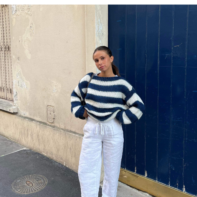 Une personne se tient sur un trottoir devant une porte bleue, vêtue dun pull rayé élégant et confortable pour toutes les saisons à rayures bleues et blanches et dun pantalon blanc chic de la boutique Olivia. Le décor respire le charme rustique avec son mur patiné.