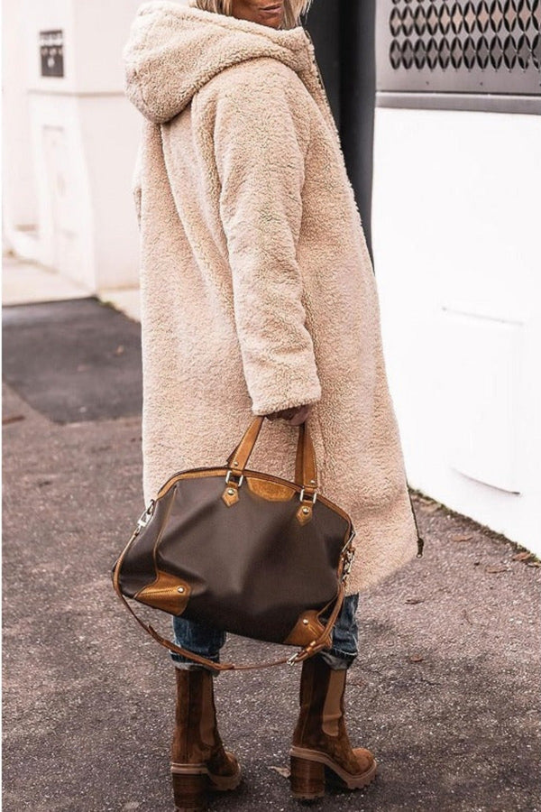 Une personne porte le Manteau réversible élégant pour femme d'Olivia's Boutique, un long manteau beige en peluche avec une capuche qui met en valeur l'élégance et la fonctionnalité, tient un grand sac marron. Des bottes marron complètent son look alors qu'elle se tient sur un trottoir urbain. En arrière-plan, il y a un bâtiment blanc avec une fenêtre à motifs.