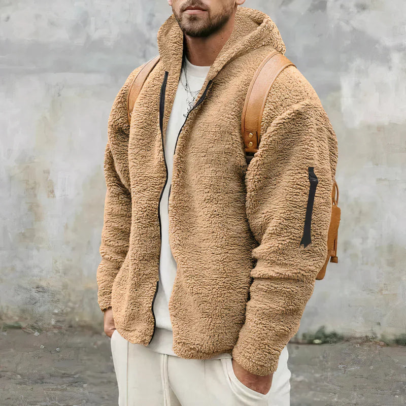 Une personne porte une élégante veste Sherpa de la boutique Olivia's en beige, associée à un pantalon crème et à un sac à dos marron, debout devant un mur gris texturé. Cette veste d'hiver confortable, officiellement nommée Veste Sherpa élégante et chaude pour l'hiver, est dotée d'un devant zippé et d'une capuche. La tête n'est pas visible sur l'image.