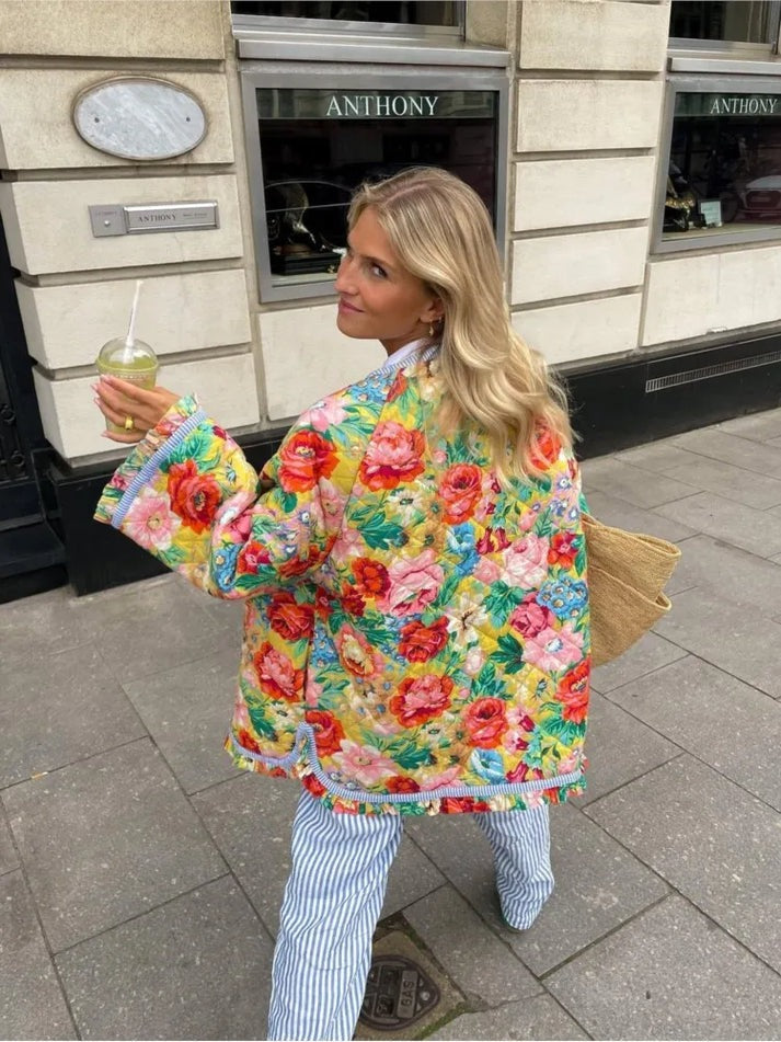 Une femme aux longs cheveux blonds déambule dans une rue de la ville, un verre à la main. Elle porte la Veste élégante avec détails en dentelle de la boutique Olivia, confectionnée avec des matériaux de qualité supérieure, associée à un pantalon rayé et porte un grand sac tissé. Le bâtiment derrière elle affiche un panneau sur lequel est écrit Anthony.