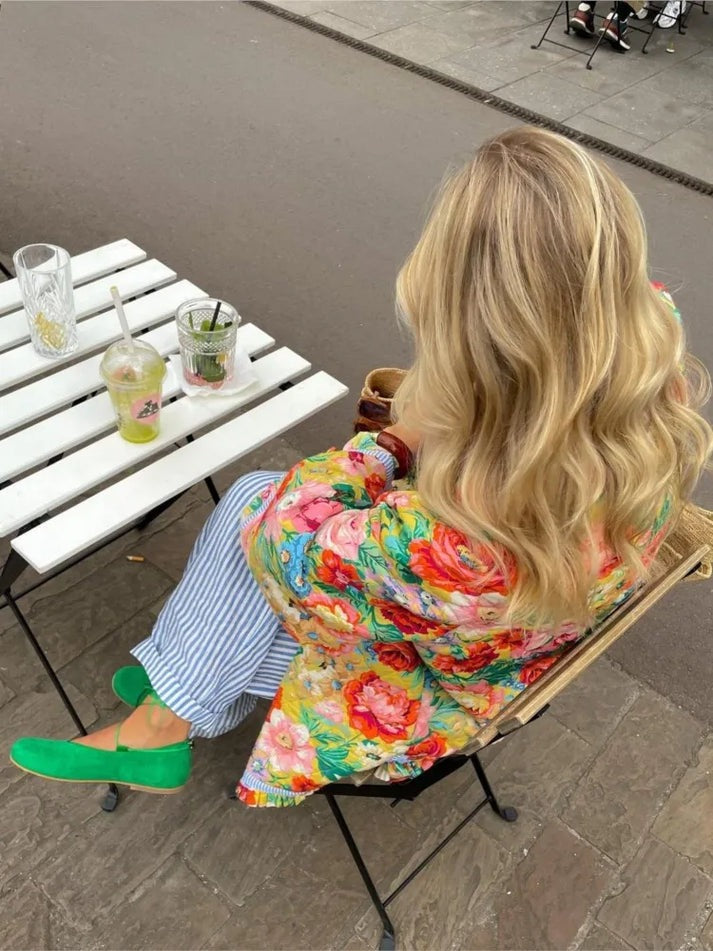 Une personne aux cheveux blonds longs est assise à une table en plein air, vêtue de la Veste élégante avec détails en dentelle – Chic et polyvalente pour toutes les occasions d'Olivia's Boutique, associée à un pantalon rayé et à des chaussures vertes. La table, située sur un trottoir, peut accueillir deux boissons, tandis qu'une route et des sièges supplémentaires sont visibles en arrière-plan.