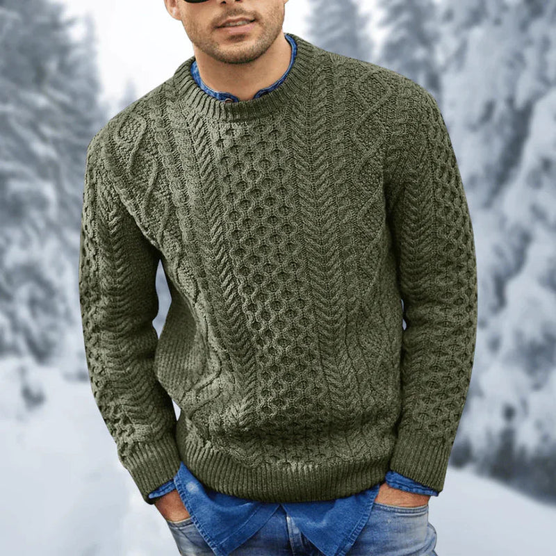 Une personne porte le pull Branimir - pull élégant avec de longues oreilles de la boutique Olivia's, debout à l'extérieur dans une forêt enneigée. Le pull en tricot vert, connu pour ses motifs torsadés complexes et sa coupe confortable, est associé à une chemise en jean bleu. En arrière-plan, des arbres flous sont magnifiquement recouverts de neige.