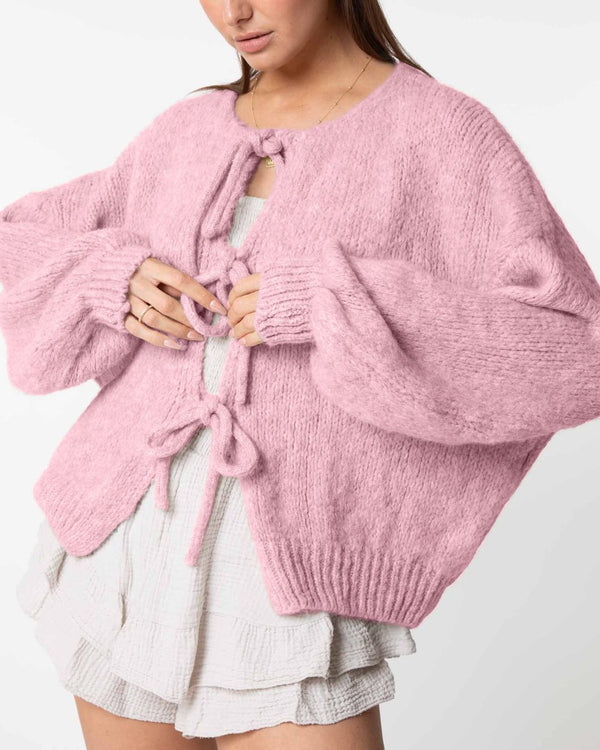 Une personne porte le cardigan oversize avec nœuds d'Olivia's Boutique en rose pastel, avec de larges manches et des fermetures à nouer, sur une jupe blanche texturée à volants. L'image met en valeur le haut du corps et les mains ajustant les liens du cardigan, capturant un style décontracté et chic.