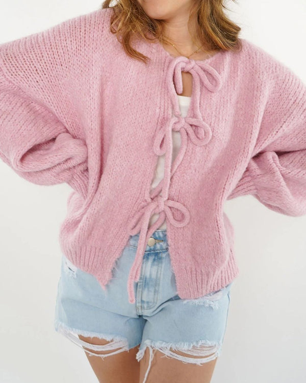 Une personne porte le cardigan oversize à nœuds d'Olivia's Boutique, dans une teinte rose pastel, sur un haut blanc. Il est associé sans effort à un short en jean bleu clair vieilli pour un look décontracté-chic sur un fond blanc uni.