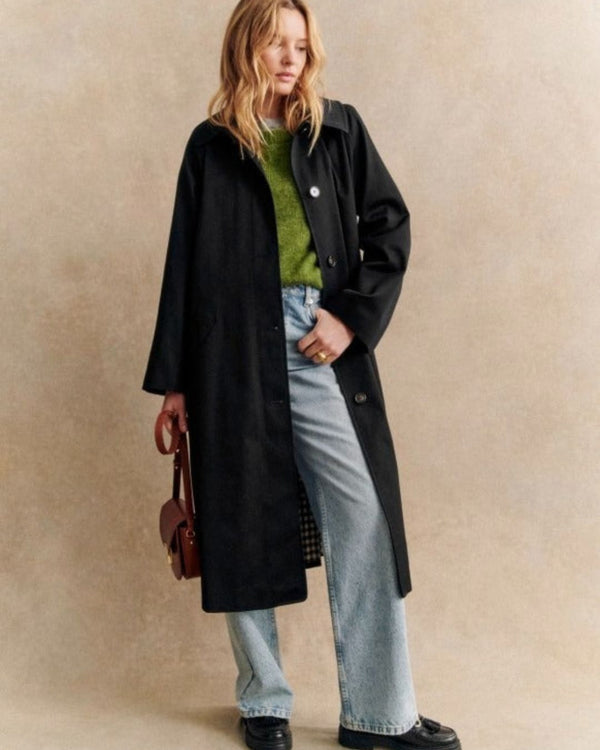 Une personne aux cheveux longs porte un trench-coat noir d'Olivia's Boutique sur un pull vert et un jean bleu à jambe large, tenant gracieusement un sac à main marron. Son élégance naturelle est mise en valeur sur le fond neutre.