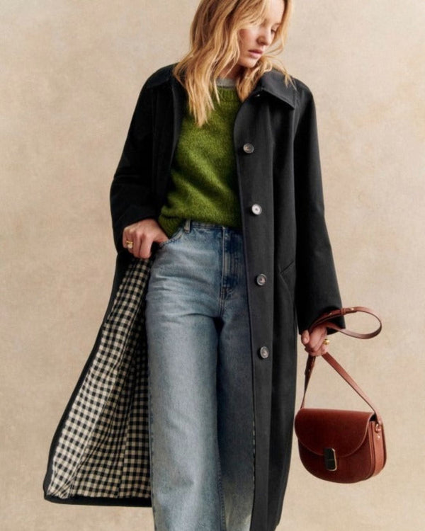 Une personne vêtue d'un trench-coat Olivia's Boutique, avec un long design noir et une doublure à carreaux, l'associe à un pull vert et un jean bleu, debout devant un fond neutre. Elle tient un sac à main marron et incline légèrement la tête vers le bas, dégageant un air d'élégance simple.