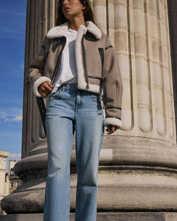 Une personne portant une veste Sherpa beige de la boutique Olivia, un t-shirt blanc et un jean bleu clair se tient devant une grande colonne classique. Le style moderne se marie avec le ciel bleu clair en arrière-plan.