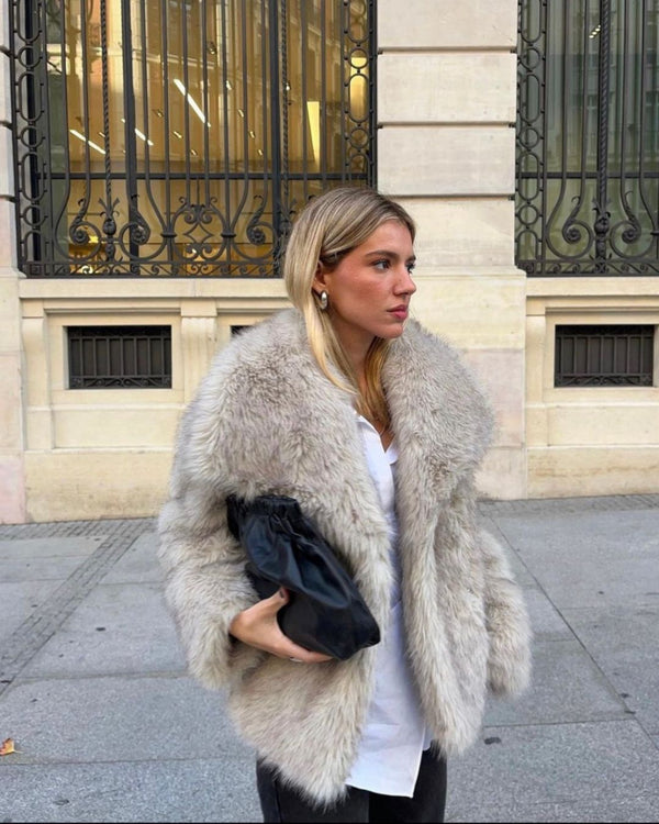 Une personne aux cheveux longs porte le Manteau fourrure tendance d'Olivia's Boutique, un manteau de fourrure moelleux, et porte un sac noir. Ils se tiennent sur un trottoir devant un bâtiment aux grandes fenêtres et aux barreaux en fer forgé ornés, incarnant le style moderne dans ce cadre urbain.