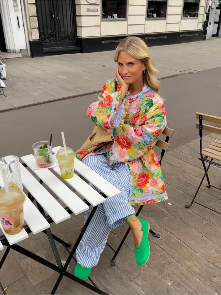 Une personne aux longs cheveux blonds est assise à une table de café en plein air, vêtue d'une veste élégante avec détails en dentelle de la boutique Olivia, complétée par un pantalon à rayures bleues et blanches et des chaussures vertes. Diverses boissons ornent la table dans un décor de rue charmant avec des bâtiments en arrière-plan.