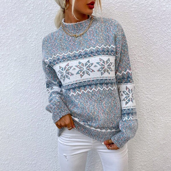 Une personne porte un Pull de Noël Motif Flocon de Neige de la boutique Olivia, avec un design bleu clair et blanc, debout contre un mur blanc, dégageant une sensation de chaleur hivernale douillette. Elle porte également un pantalon blanc et un collier doré, les cheveux attachés en arrière et les mains nonchalamment glissées dans leurs poches.