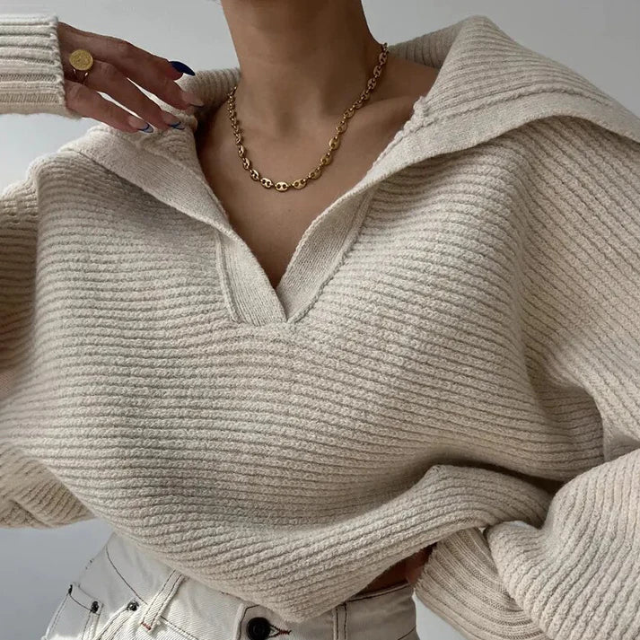 Une personne porte le Pull en gros tricot élégant pour les journées froides de la marque Olivia's Boutique en beige, associé à un jean blanc. Elle porte un collier en chaîne dorée et sa main est levée près de l'encolure, révélant une bague à un doigt. Le design moderne de la tenue est accentué par l'aspect luxueux du tissu doux.