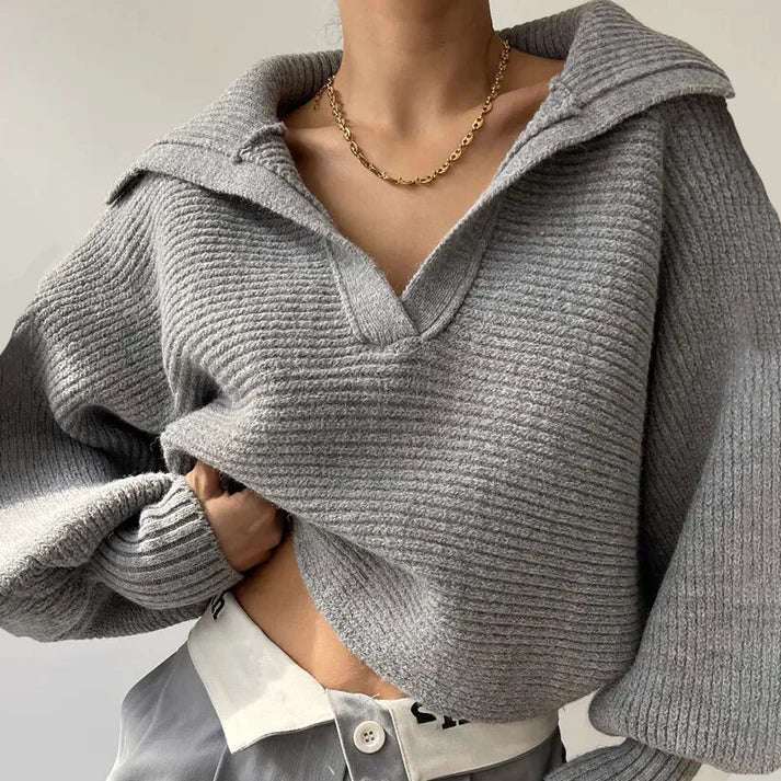 Une personne porte le Pull en gros tricot élégant pour les journées froides d'Olivia's Boutique en tricot gris texturé avec un large col sur une chemise blanche et un pantalon gris. Fabriqué en tissu doux, le pull a une coupe légèrement oversize pour une ambiance décontractée. Ils accessoirisent avec un simple collier en chaîne dorée sur le fond neutre minimaliste.