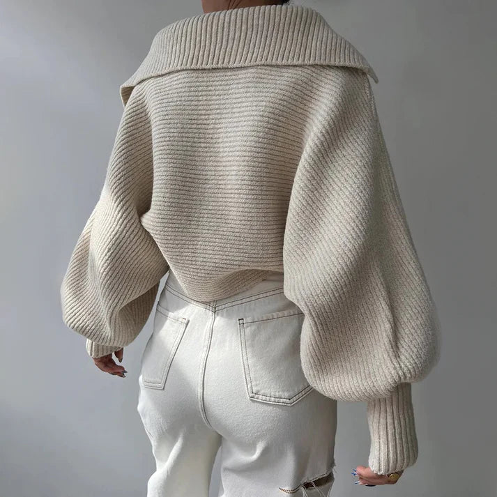 On voit une personne portant le pull en gros tricot élégant pour les journées froides d'Olivia's Boutique, avec une coupe ample et des manches surdimensionnées dans une teinte crème, associé à un pantalon blanc à taille haute. L'image capture la vue arrière, mettant en valeur le tissu doux du pull et son style contemporain.