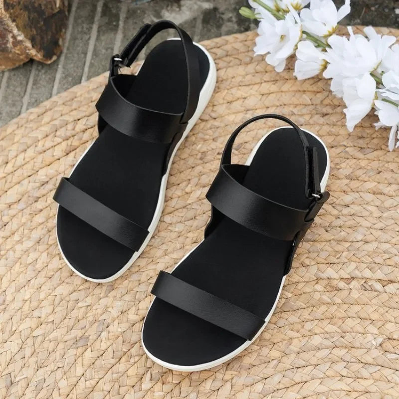 Une paire de Sandales d'été orthopédiques d'Olivia's Boutique à semelles blanches repose sur un tapis tissé, accentué de fleurs blanches. Conçues pour une tenue décontractée, ces sandales noires offrent un maintien optimal avec une conception à bride qui assure à la fois style et confort.