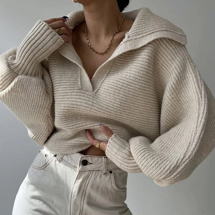 Une personne est vêtue du Pull en gros tricot élégant pour les journées froides d'Olivia's Boutique, de couleur crème et surdimensionné avec un grand col. Elle l'associe à un pantalon blanc taille haute et accessoirise avec un collier en chaîne dorée et des bagues, se tenant sur un fond neutre, dégageant un air de design moderne.