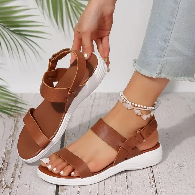 Vêtue d'un jean bleu clair, une personne ajuste la bride d'une sandale d'été orthopédique Olivia's Boutique de couleur marron à semelles blanches. Des bracelets de cheville à perles blanches promettent du confort, tandis que des frondes vertes ornent le fond en bois.