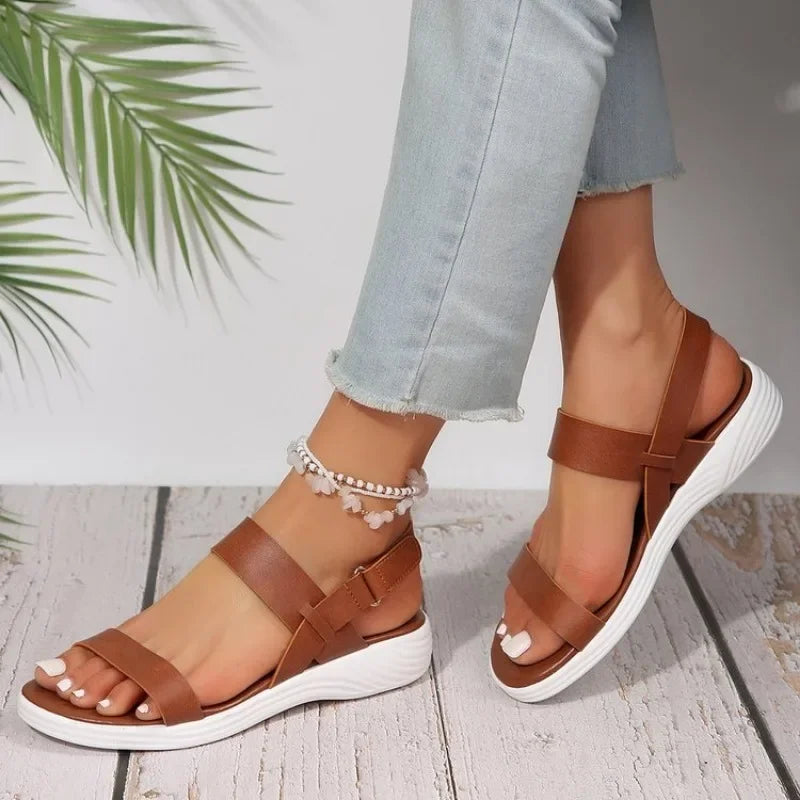 Les pieds dans la boutique Olivia Sandales d'été orthopédiques, lanières marron et semelles blanches, associées à un jean bleu clair retroussé. Une chevillère perlée orne une cheville sur fond de parquet et de feuilles de palmier vertes.