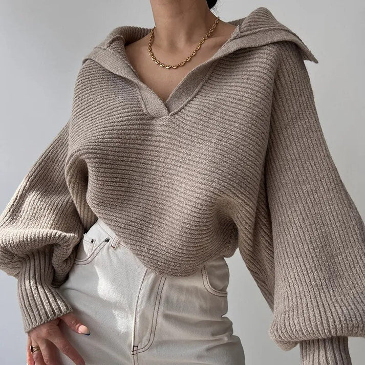 Une personne porte le pull en gros tricot élégant pour les journées froides de la boutique Olivia en beige, avec un large col et des manches ballon, associé à un pantalon blanc à taille haute. Le tissu doux du pull complète parfaitement son style, accentué par un collier en chaîne dorée et une main nonchalamment placée dans sa poche sur un fond doucement éclairé.