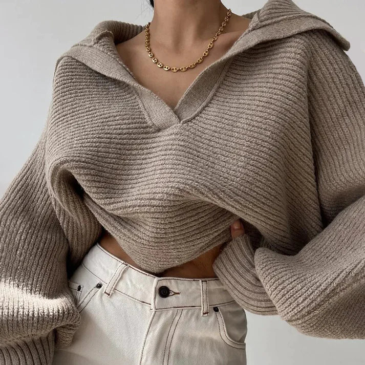 Une personne est habillée d'un pull en gros tricot élégant pour les journées froides d'Olivia's Boutique, avec un col en V et un grand col en tissu doux. Ils l'ont associé à un pantalon beige taille haute et à un simple collier en chaîne dorée, incarnant un design moderne sur fond gris uni.