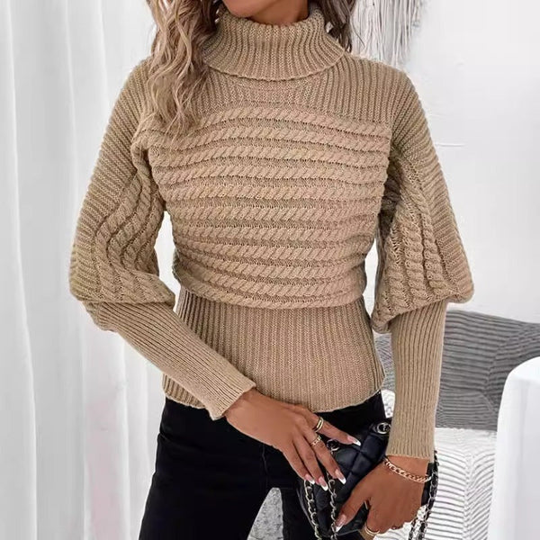 Vêtue du Pull à Col Haut en Tricot Torsadé beige d'Olivia's Boutique, associé à un pantalon noir, la personne tient un petit sac à main noir doté d'une bandoulière en chaîne. Le décor comprend des rideaux blancs et une ambiance intérieure cosy.