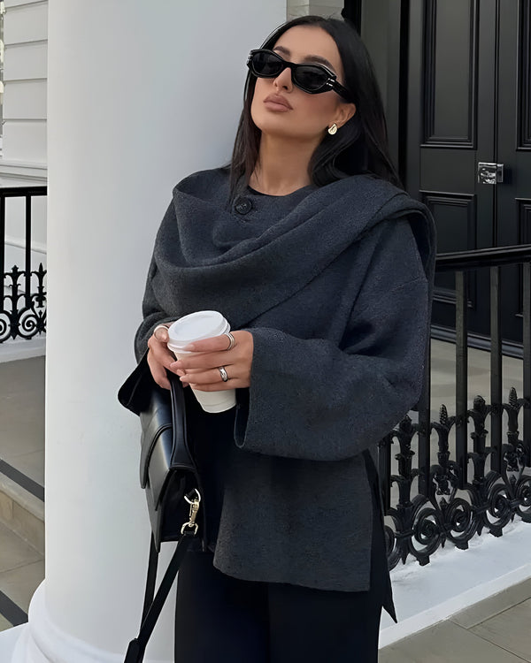 Une personne aux cheveux longs et foncés se tient dehors, portant des lunettes de soleil et un élégant Manteau Trendy à Foulard de la boutique Olivia. Elle tient une tasse de café dans une main et un sac à main noir dans l'autre, debout devant une colonne blanche avec une rampe noire derrière elle.