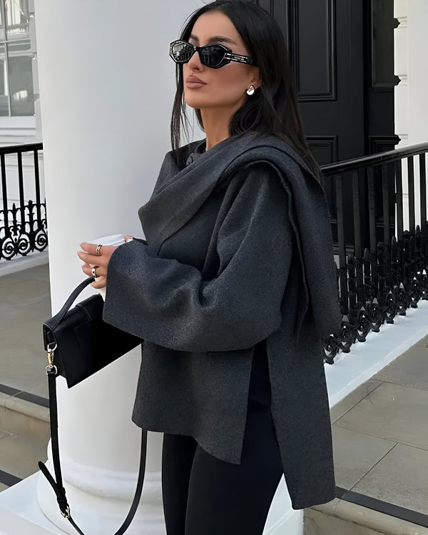 Une personne se tient dehors, reflétant l'élégance moderne dans le « Manteau Trendy à Foulard » d'Olivia's Boutique, une tenue sombre surdimensionnée avec des foulards superposés. Arborant de longs cheveux noirs et des lunettes de soleil, elle tient une tasse blanche. Un sac à main noir repose sur son épaule, se détachant sur une colonne blanche et une balustrade noire derrière elle.