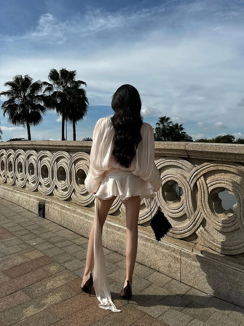 Une femme vêtue d'une robe fluide élégante et confortable pour toutes les occasions d'Olivia's Boutique et de talons hauts se tient debout sur une passerelle en pierre, surplombant une balustrade avec des palmiers et un ciel bleu en arrière-plan. Elle tient une pochette noire, mettant en valeur le confort exceptionnel de son ensemble au design intemporel.